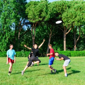 Group of mixed young teenagers people in casual wear playing with plastic flying disc game in a park oudoors. jumping woman catch a disk to a teammate in a match. milennial friends outside in a garden